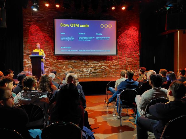 Andrea Verlicchi wearing a yellow sweater speaking on stage at the performance.sync() meetup in Amsterdam. The current slide shows "Slow GTM Code. Audit. Remove. Update. Not enough?"
